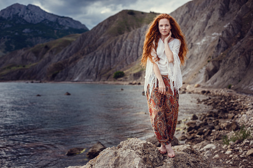 Young red-haired girl in the style of Boho. Enjoys the beauty of the sea coast. The idea and concept of freedom and hippies