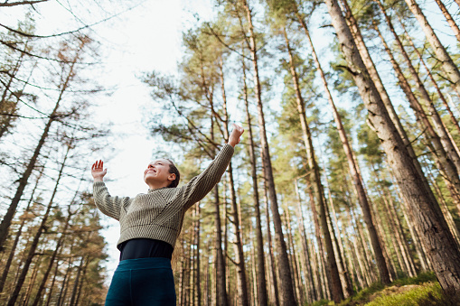 Low angle view of a teenage girl walking through Thrunton woods in the North East of England. She is on a digital detox and is practising mindfulness, holding her arms up in the air.