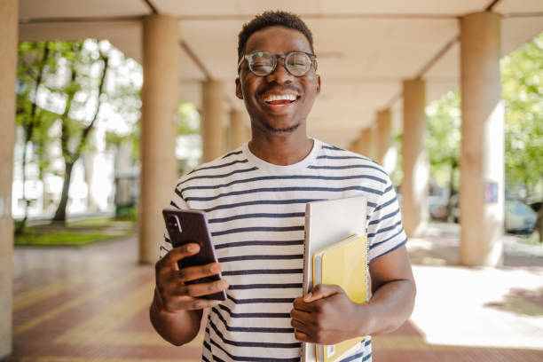 Portrait of a young confident African-American student using mobile phone on the move Portrait of an African-American student carrying a notebook and a laptop in front of the University education student mobile phone university stock pictures, royalty-free photos & images