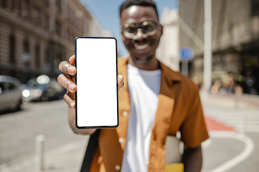 A young African-American man is on the street, he is holding a white mobile phone screen and smiling