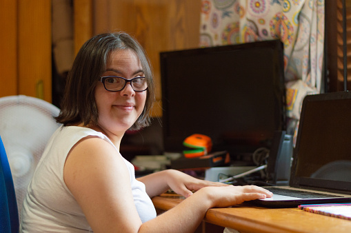 Young adult female with Down syndrome sitting in the bedroom of her home, using a laptop computer and smiling at the camera.