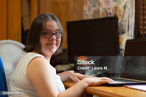 istock Young adult female with Down syndrome sitting in the bedroom of her home, using a laptop computer and smiling at the camera. 1339297278