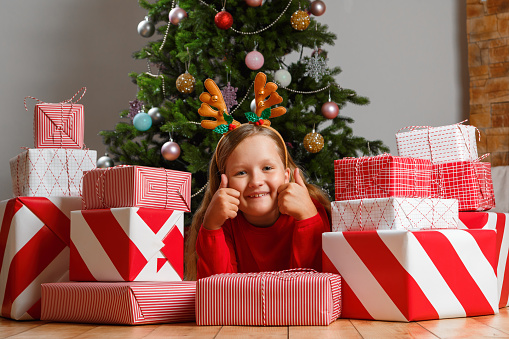 The child lies on the floor surrounded by boxes with gifts against the background of a Christmas tree. Little girl in a deer headband showing thumb up