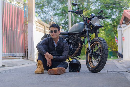 Portrait of Asian young handsome man biker wear black leather jacket and sunglasses sitting smile on concrete street road front house and look at camera with safety helmet and motorcycle in outdoors.
