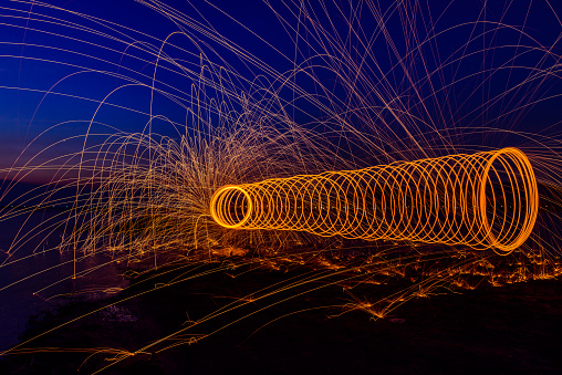 Beautiful fireworks from burning and spinning steel wool by expert at seaside in twilight on blue sky background. Abstract long exposure photography for circle fire glowing sparks by steel wool burn.