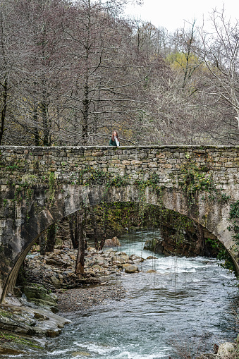 Distant view of female standing on shabby stone bridge crossing river in autumn forest on cloudy day. Rural weekend getaway concept