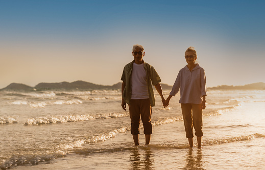 Asian senior couple tourist wear sunglasses hand in hand walking in sea water on beach in summer vacation. Happy family woman and man gray hair elderly older retire relax together while sunset seaside