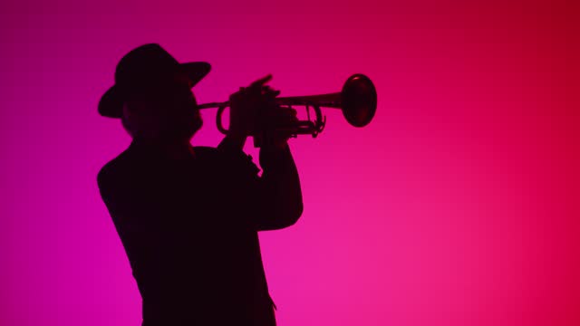 Man plays music on trumpet in room flickering with different neon lights, front view. Jazz performer in hat and suit plays trumpet