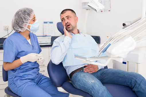 Unhappy american man in medical chair complains of toothache to female dentist at modern dental clinic