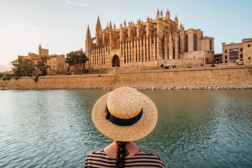 Rear view of a woman with a straw hat while she's admiring the Cathedral de Santa María de Palma de Mallorca at sunset. The Cathedral reflects in the water.