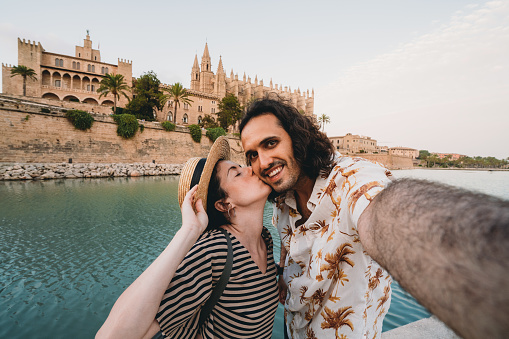 A couple is taking a selfie near the Cathedral de Palma de Mallorca at sunset. Pov view. The woman is kissing the man.