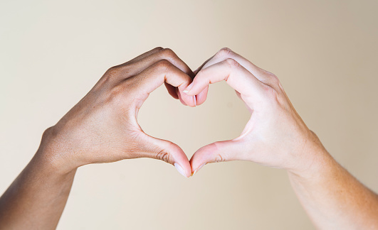 African hand and Caucasian hand make heart, beige background