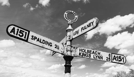 An old fashioned British road sign on the A151 pointing to Spalding, Holbeach, Kings Lynn and Low Fulney Estate in black and white in front of clouds