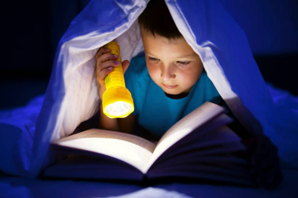 istock A young boy reading a book under the covers with a flashlight at dark night time. 1339326706