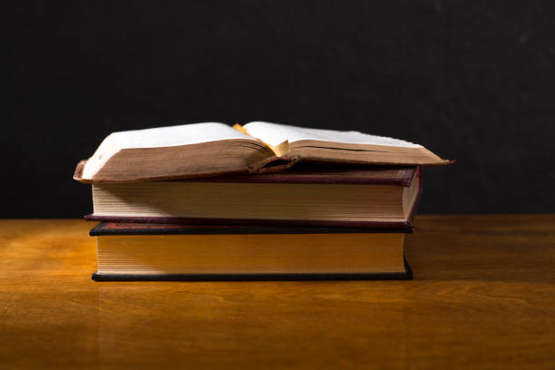 istock Open  book or Bible on stack of other leather bound books.  Highly polished wood table.  Black background.  Room for copy. 1339327457