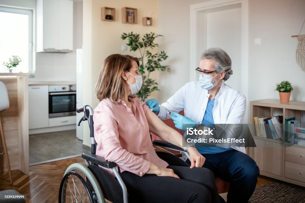Vaccination for Homebound People Covid-19 vaccination for homebound people. Doctor giving coronavirus vaccine to a handicapped female patient in a wheelchair in the living room Male Nurse Stock Photo
