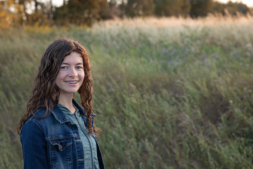 Portrait of a teen girl with long brown curly hair, smiling, with braces, and looking at camera, standing in an agricultural field in sunset in autumn
