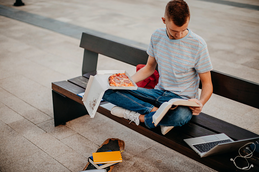 College student sitting outside on bench eating pizza while reading