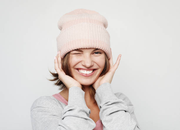 Funny young woman in a pink beanie hat makes faces and poses over light grey background stock photo