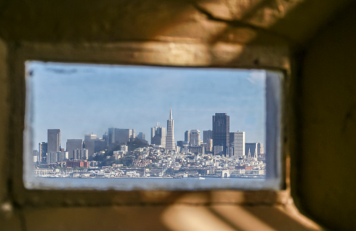 USA, California, San Francisco - May 6, 2007: Alcatraz. City skyline visible through small thick glass prison window under blue sky. Faded brown wall as frame.