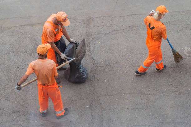 Krasnoyarsk, Russia - 19 August, 2021: Utilities workers in orange uniforms with scoops and brooms sweep the sidewalk and put trash in a black plastic bag