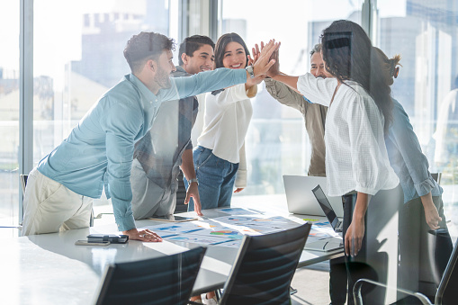Business team celebrating success with a high five. They are casually dressed in a board room with a window behind them. There is a diverse mix of ethnicities and ages