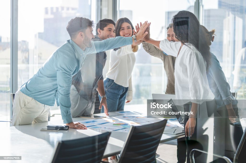 Business team celebrating success with a high five. Business team celebrating success with a high five. They are casually dressed in a board room with a window behind them. There is a diverse mix of ethnicities and ages Aspirations Stock Photo