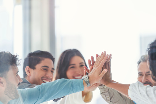 Business team celebrating success with a high five. They are casually dressed in a board room with a window behind them. There is a diverse mix of ethnicities and ages