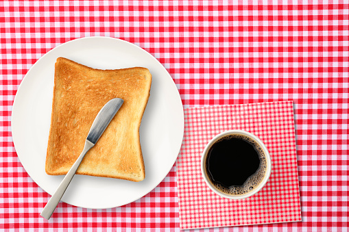 Overhead shot of slice toasted bread and butter knife on a white plate with a cup of coffee, on a gingham check tablecloth.