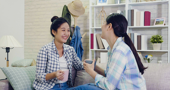 Two chinese girls sisters sitting on sofa have fun laughing and chatting together at home. young asian women talking joyfully holding cups drinking coffee indoors. best friends spending time together