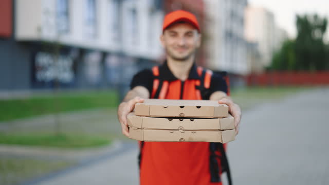 Attractive young delivery man courier in red cap smiling into camera holding pizza boxes delivering fast food around city. Outdoor portrait. Urban worker. Concept of courier, home delivery, pizza