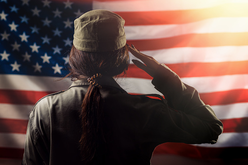 Veterans Day, Memorial Day, Independence Day. A female soldier saluting against the background of the American flag. Back view. Light. Concept of national American holidays and patriotism.