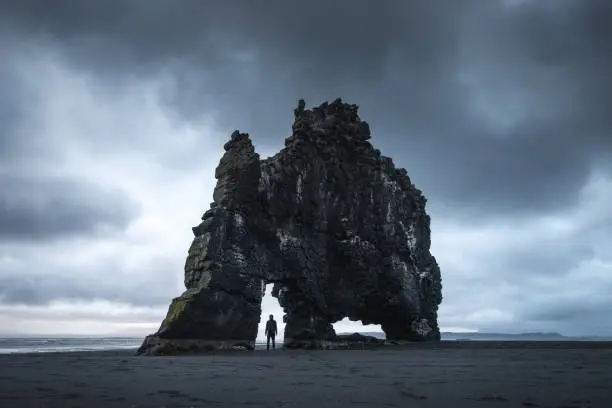 Man standing under the famous rock Hvitserkur on the black beach in north Iceland.