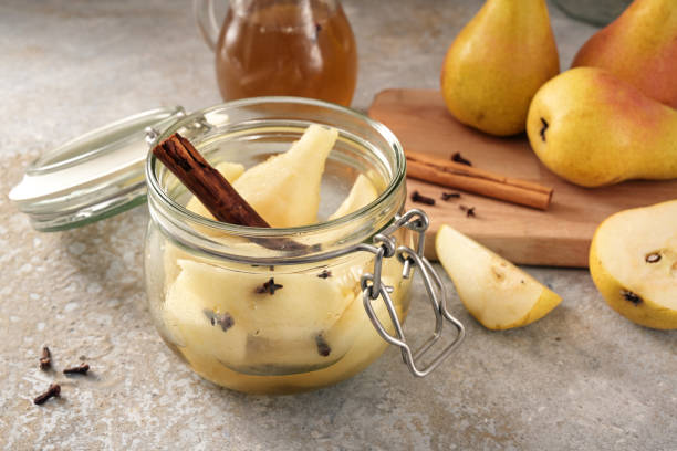 Canning pears with cinnamon and cloves in a glass jar, and some fresh fruits on a cutting board, rustic gray stone background, selected focus stock photo