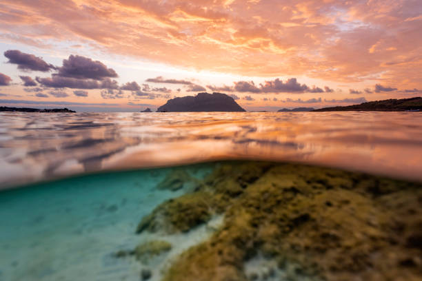 (Selective focus) Split shot, over under water surface. Defocused waves in the foreground with Tavolara Island on the surface during a dramatic sunrise. Porto Istana, Sardinia, Italy. (Selective focus) Split shot, over under water surface. Defocused waves in the foreground with Tavolara Island on the surface during a dramatic sunrise. Porto Istana, Sardinia, Italy. istana stock pictures, royalty-free photos & images