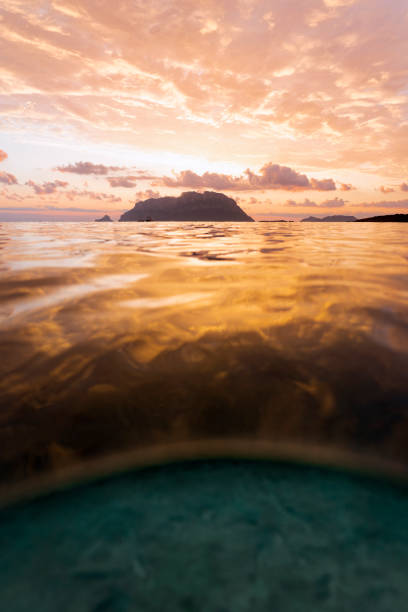 (Selective focus) Split shot, over under water surface. Defocused waves in the foreground with Tavolara Island on the surface during a dramatic sunrise. Porto Istana, Sardinia, Italy. (Selective focus) Split shot, over under water surface. Defocused waves in the foreground with Tavolara Island on the surface during a dramatic sunrise. Porto Istana, Sardinia, Italy. istana stock pictures, royalty-free photos & images