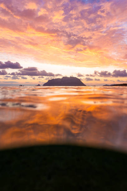 (Selective focus) Split shot, over under water surface. Defocused waves in the foreground with Tavolara Island on the surface during a dramatic sunrise. Porto Istana, Sardinia, Italy. (Selective focus) Split shot, over under water surface. Defocused waves in the foreground with Tavolara Island on the surface during a dramatic sunrise. Porto Istana, Sardinia, Italy. istana stock pictures, royalty-free photos & images