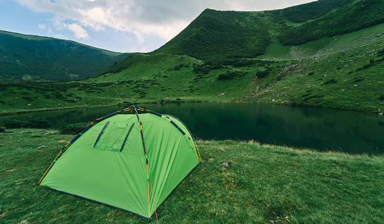A tent by the lake at the foot of green mountains covered with bushes and grass in cloudy summer weather. Tourism, rest, recreation. Mountain landscape.