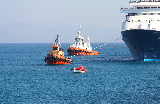 Tugboats and a pilot boat maneuvering a large tourist ferry into a harbour on the Greek island of Rhodes. It’s a hot, summer day with blue, cloudless sky. Mountains are visible on the horizon. Dominant colors: blue, red, orange, black, white.