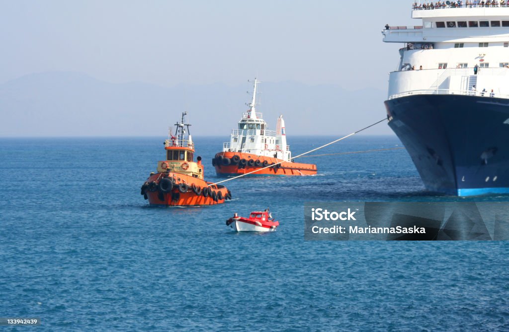 Tugboats towing a ferry into a harbor Tugboats and a pilot boat maneuvering a large tourist ferry into a harbour on the Greek island of Rhodes. It’s a hot, summer day with blue, cloudless sky. Mountains are visible on the horizon. Dominant colors: blue, red, orange, black, white. Tugboat Stock Photo