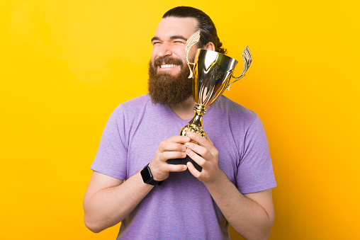 Happy excited bearded man is embracing his golden trophy over yellow background.
