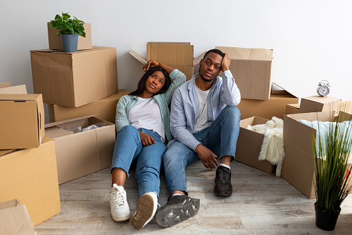 Sad black spouses exhausted after moving to new apartment, sitting among cardboard boxes and resting, having problems during relocation to own home