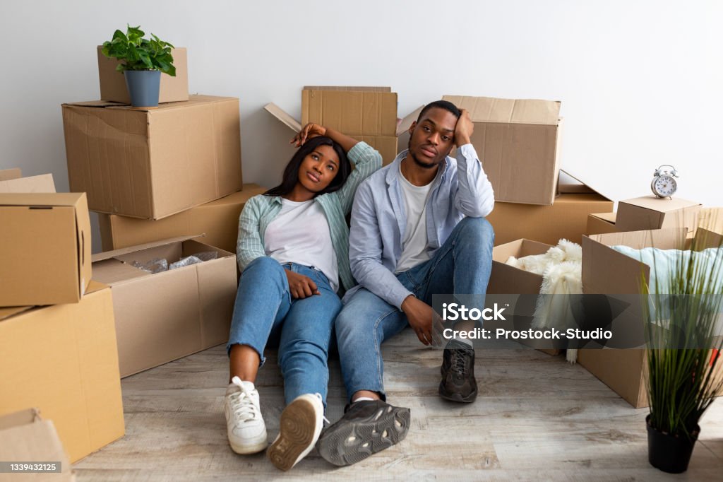 Sad black spouses exhausted after moving to new apartment, sitting among cardboard boxes and resting Sad black spouses exhausted after moving to new apartment, sitting among cardboard boxes and resting, having problems during relocation to own home Relocation Stock Photo