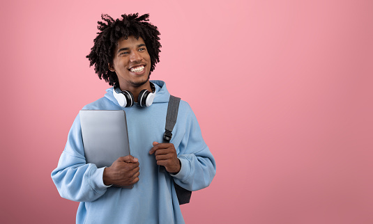 Estudiante adolescente afroamericano positivo con mochila, tablet PC y auriculares estudiando en línea sobre fondo rosa photo