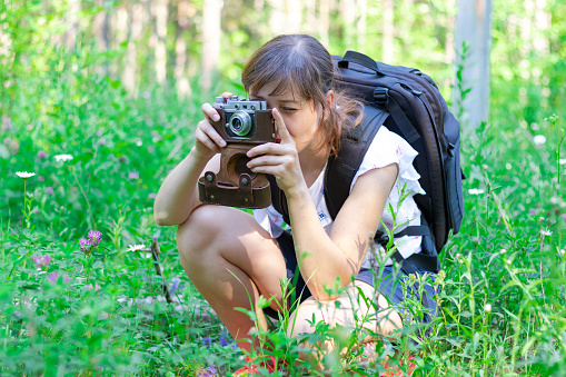 Young girl tourist with a backpack and a camera in the forest on a background of green grass on a summer sunny day. Selective focus