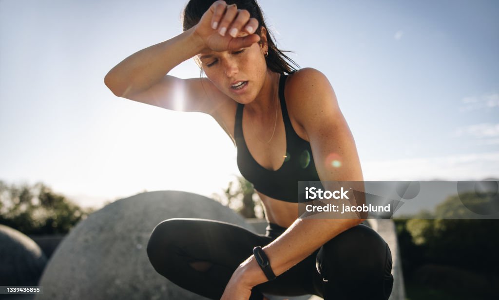 Tired woman sitting and resting after workout Tired woman sitting and resting after workout. Woman feeling exhausted after training session wiping her sweat from her forehead. Sweat Stock Photo