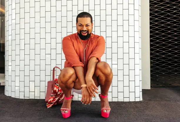 Excited drag queen smiling at the camera cheerfully while squatting against a white background. Non-conforming young man wearing make up and jewellery. Young man embracing his queer identity.