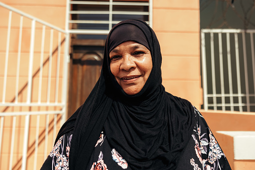 Cheerful Muslim woman smiling at the camera while standing in front of her home during the day. Self-confident middle aged woman wearing a hijab and standing alone.