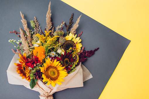 Fall bouquet of yellow red orange flowers wrapped in paper and arranged on grey and yellow background. Sunflowers, amaranth, daisies with zinnias and grasses