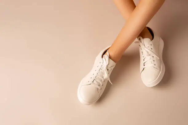Casual footwear - white leather sneakers shoes and girlâs crossed legs on nude background. Layout with free copy (text) space.